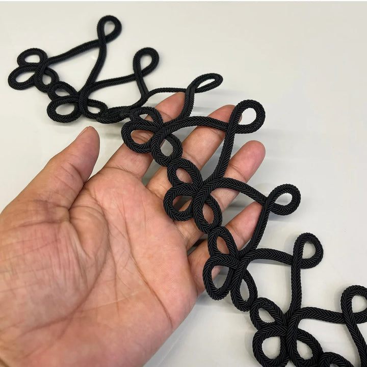 A hand holding a black braided decorative cord trim with repeating looped knot patterns, shown against a plain light background.