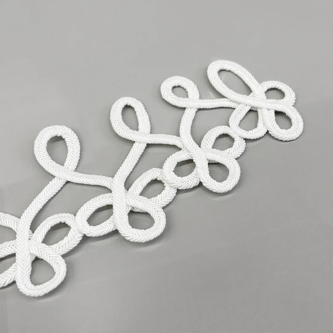 White braided rope decorative trim with an elegant looped pattern, laid diagonally on a smooth gray surface.