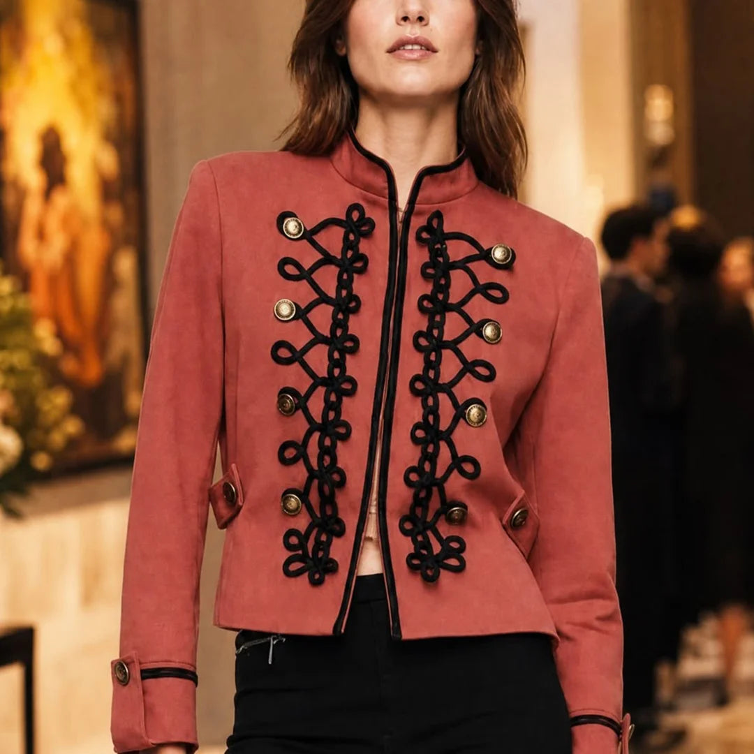 A woman wearing a cropped dusty rose jacket with ornate black decorative trim and gold-tone buttons, paired with black pants in an indoor setting.