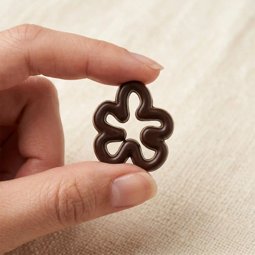 ANI Accessories dark brown five-petal flower charm held between fingertips to show real size, on beige linen background