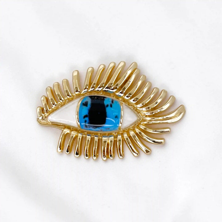 ANI Accessories gold evil eye brooch with blue stone, white enamel and long curled lashes on white satin with white color background 