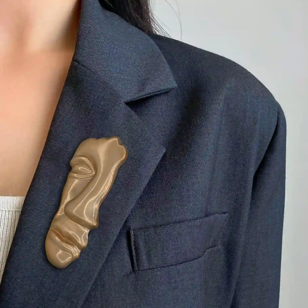 ANI Accessories matte gold face brooch on the lapel of a grey blazer 