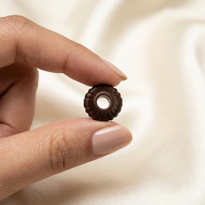 ANI Accessories mini dark brown fluted cord end bead held between fingertips, top view of centre hole on cream satin 
Background 